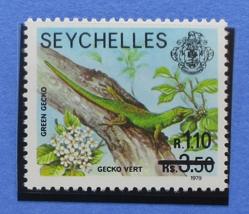 A vintage Seychelles postage stamp featuring a green gecko on a tree branch with white flowers, printed on a light blue background, shown against a blue backdrop.