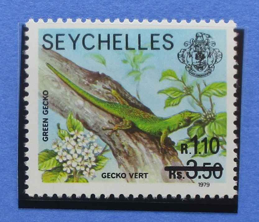 A vintage Seychelles postage stamp featuring a green gecko on a tree branch with white flowers, printed on a light blue background, shown against a blue backdrop.