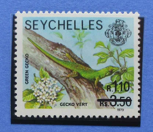 A vintage Seychelles postage stamp featuring a green gecko on a tree branch with white flowers, printed on a light blue background, shown against a blue backdrop.