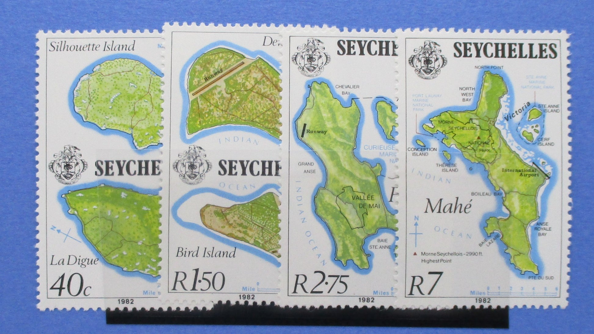 Four vintage Seychelles postage stamps displayed side by side on a blue background. The stamps feature illustrated maps of Seychelles islands, including La Digue, Bird Island, and Mahé, with denominations 40c, R1.50, R2.75, R7