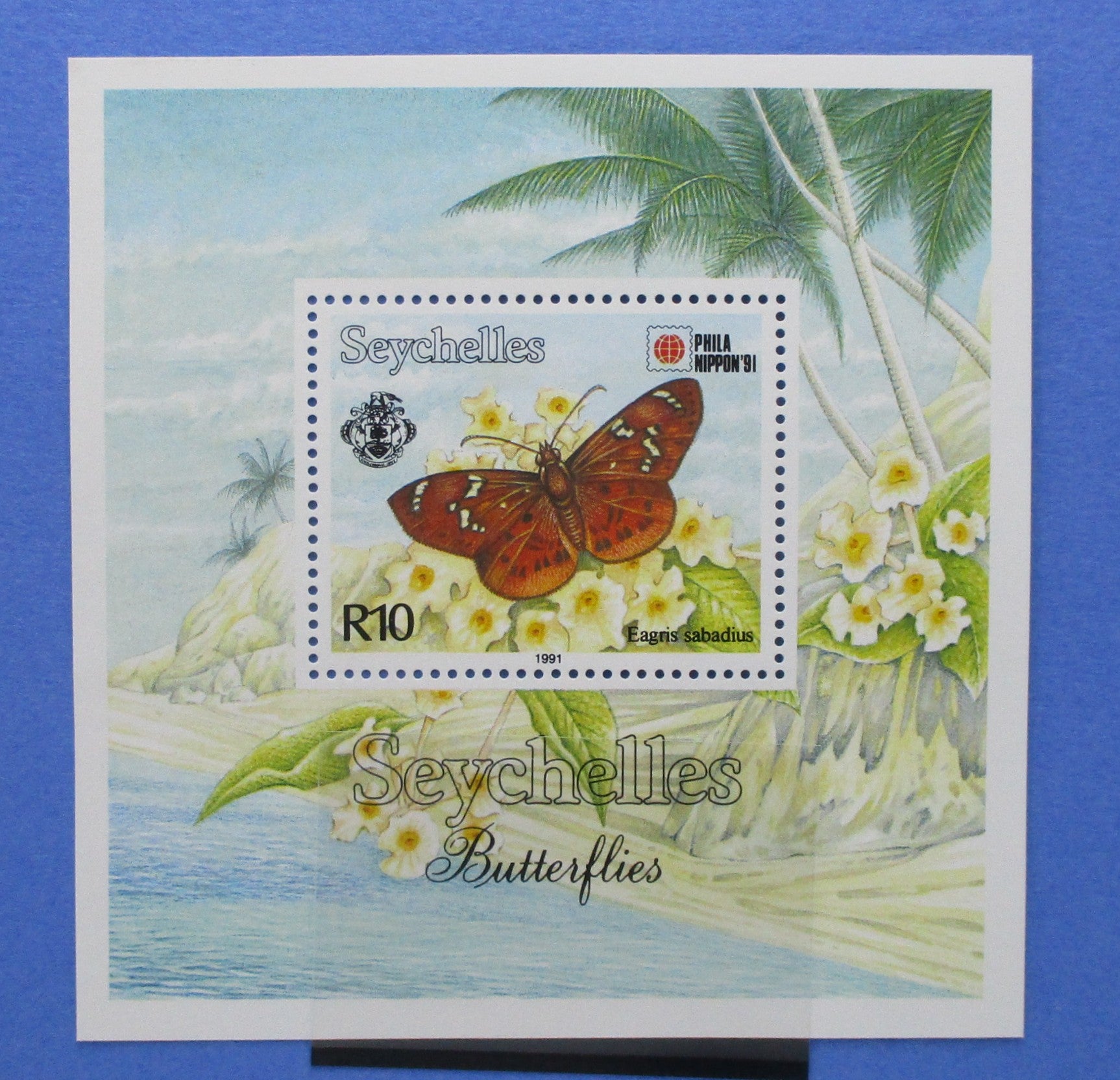 Seychelles postage stamp souvenir sheet featuring a butterfly illustration over a tropical beach scene with palm trees and white flowers, displayed against a blue background.