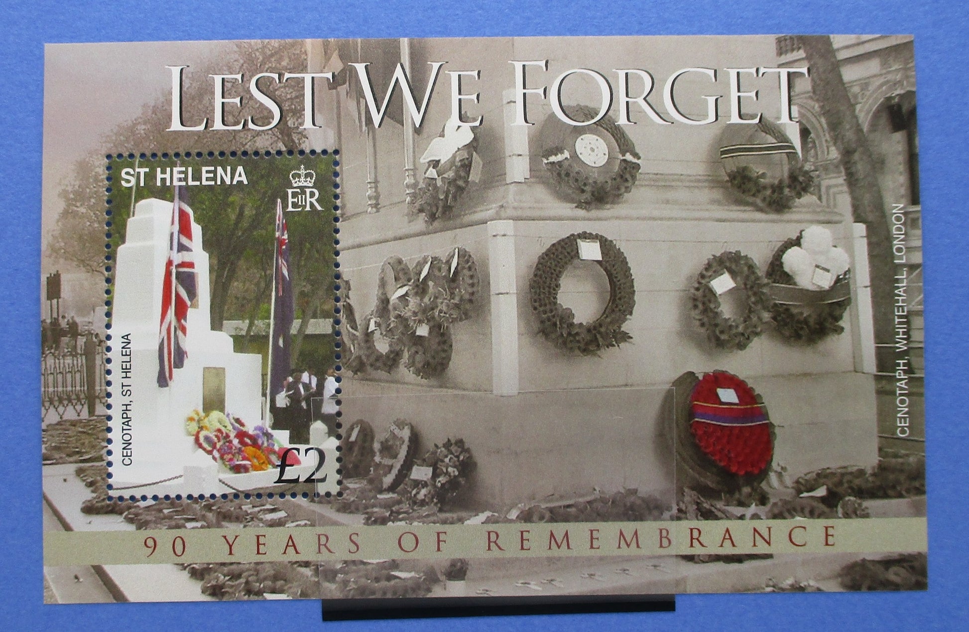A commemorative St Helena postage stamp sheet titled 'Lest We Forget' with a memorial scene, wreaths, and a featured stamp on the left. The sheet is photographed against a blue background.